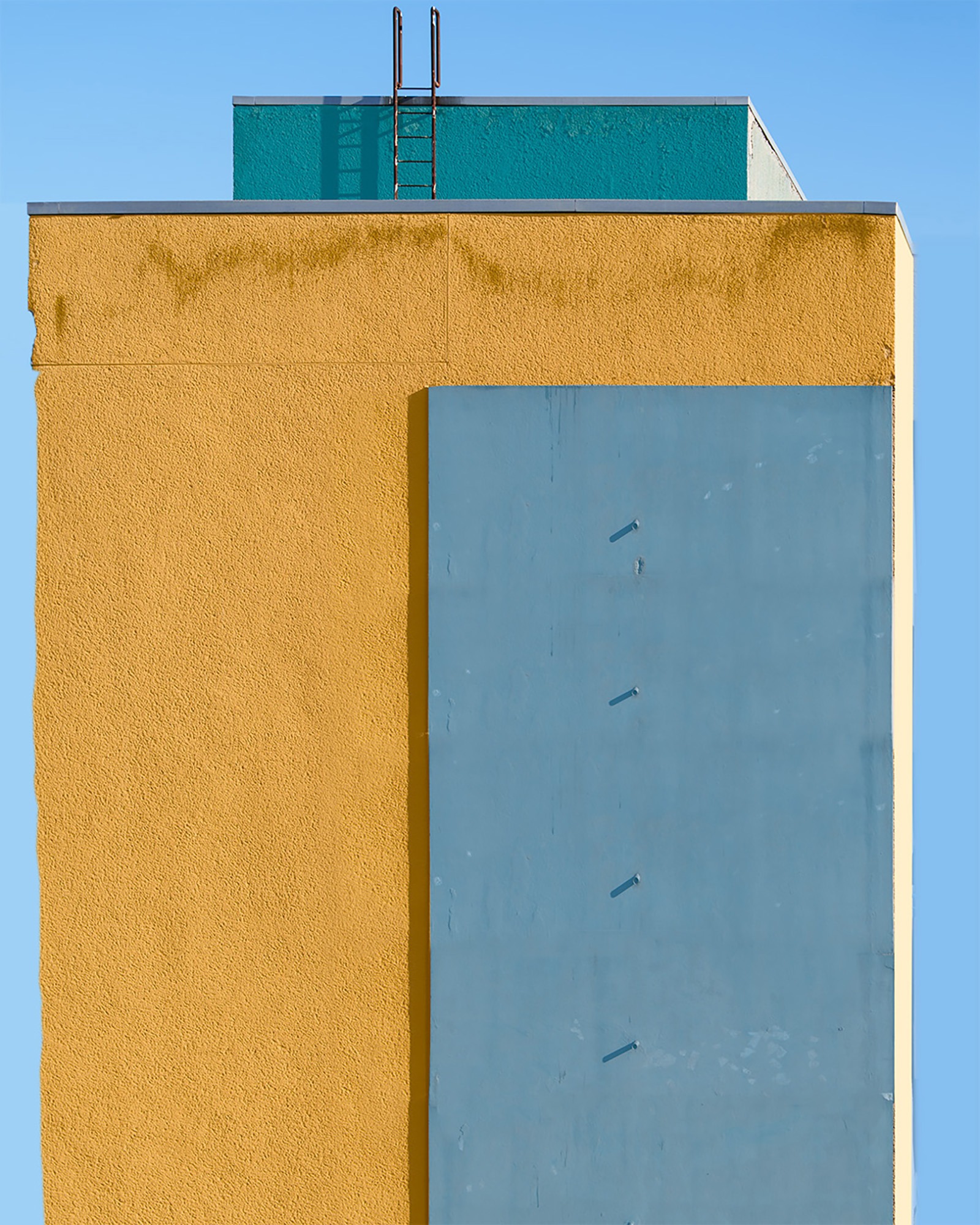 Colour-shifted fa&ccedil;ade study in Berlin-Kreuzberg: an orange rendered block seen from the corner with a large teal panel set into it, four short pipe stubs in a line, and a teal roof box with a rusted ladder against a blue sky.