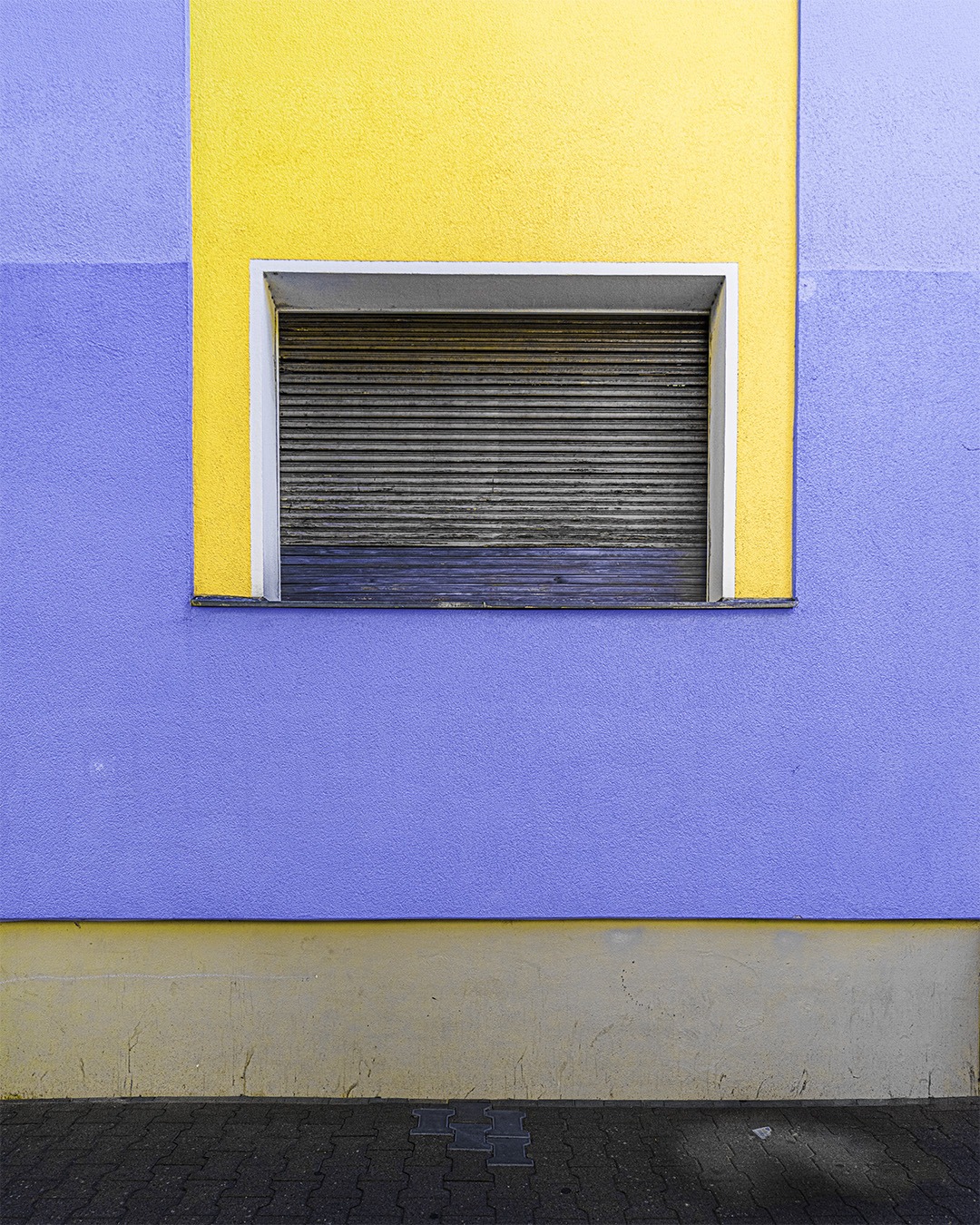 Blue rendered wall with a yellow painted frame around a single window, the roller shutter fully down, a yellow band and paving below