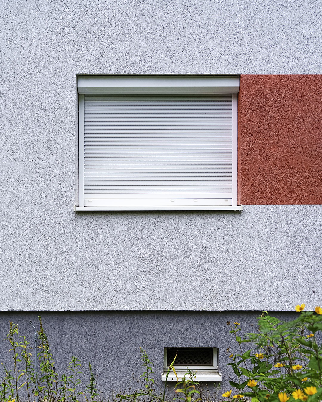 Pale grey-lilac wall with a closed white roller shutter beside a red painted rectangle, a blue-grey plinth with a basement vent and a strip of flowering plants below