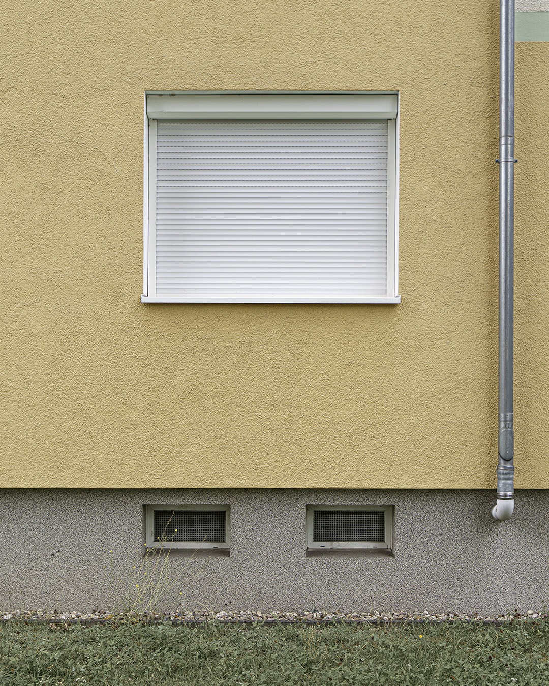 Ochre rendered wall with a closed white roller shutter, a downpipe at the right edge, a grey plinth with two basement vents and a strip of grass
