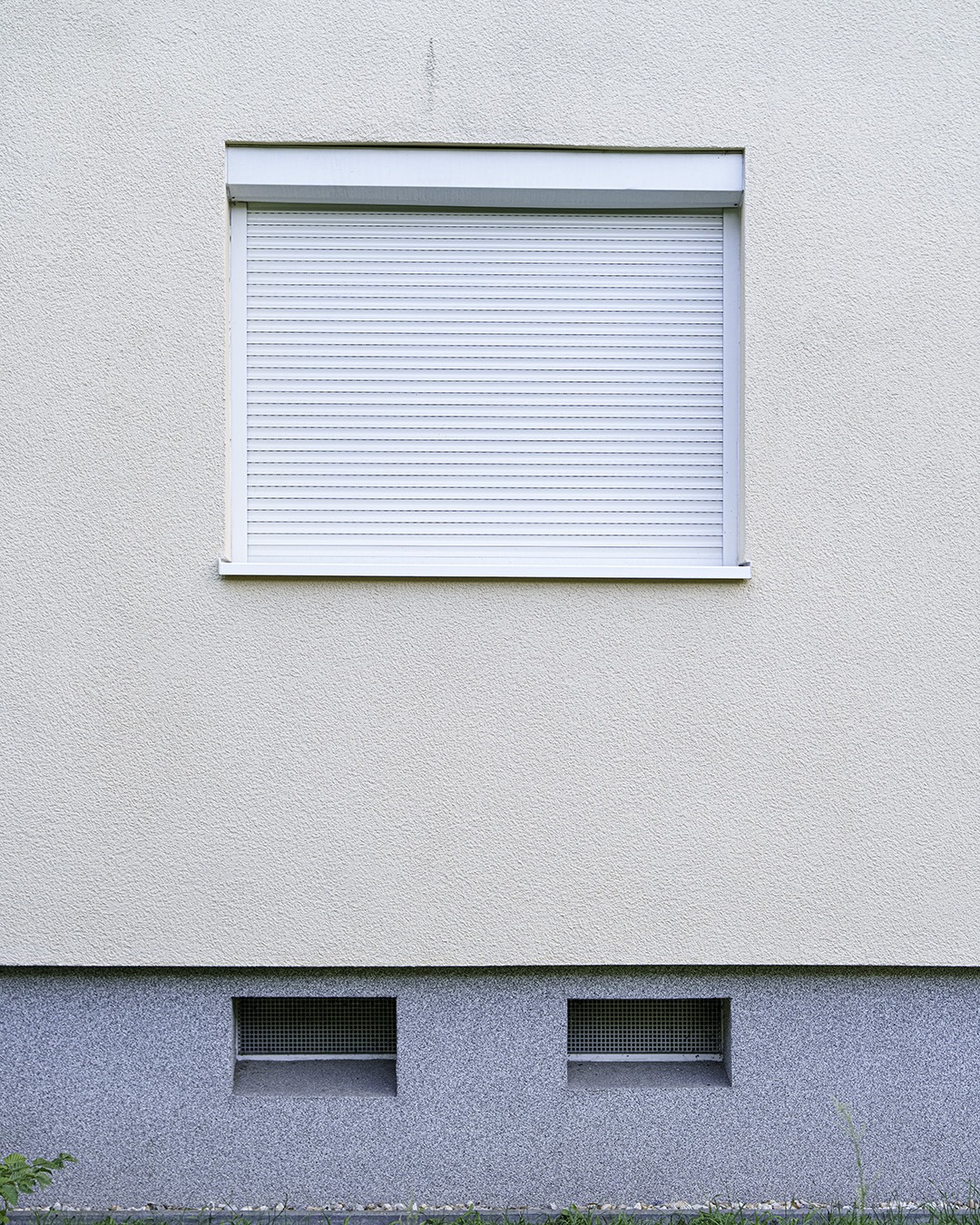Almost white wall with a closed white roller shutter, a blue-grey plinth with two basement vents and a strip of grass