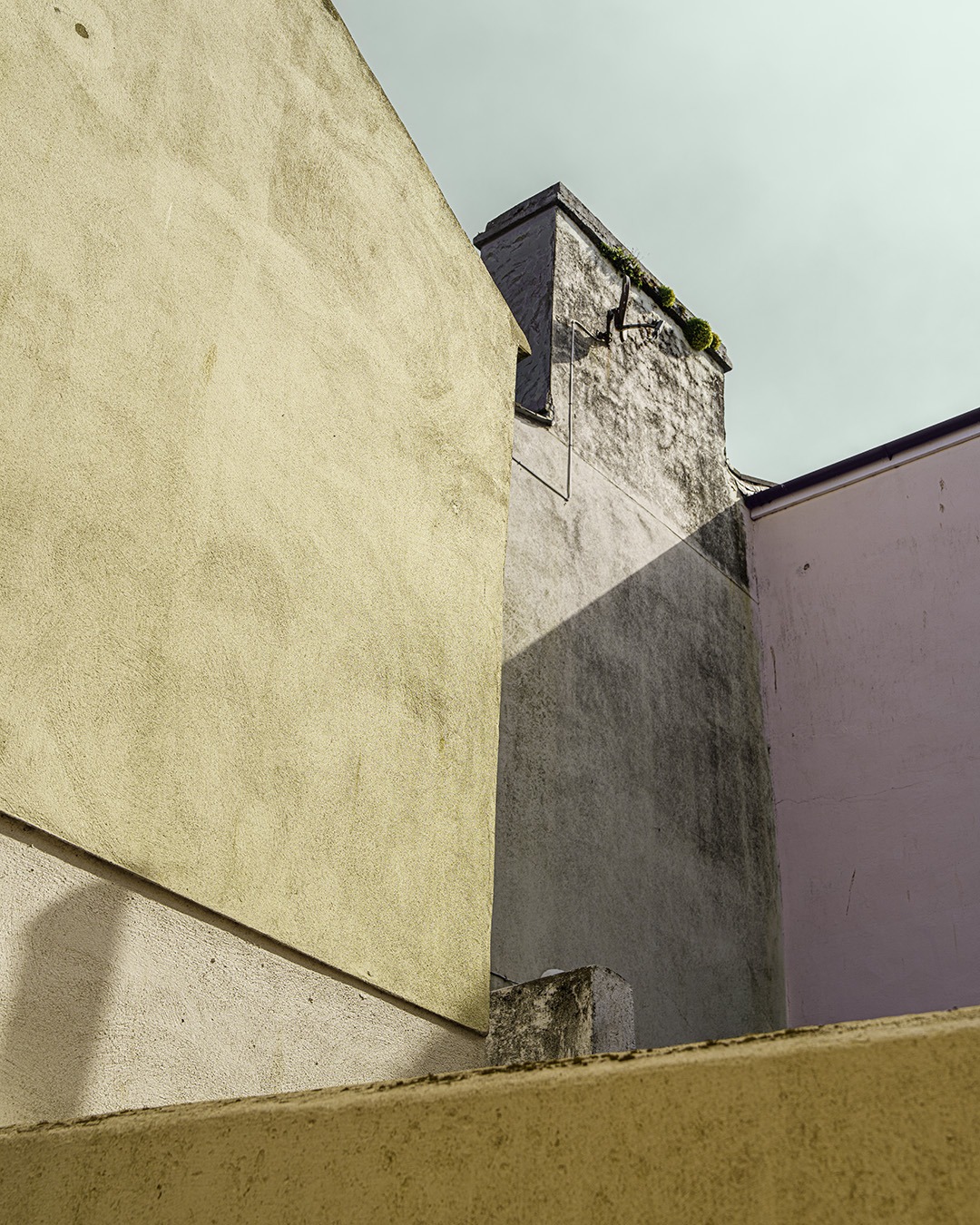 Colour-shifted fa&ccedil;ade study in Douglas, Isle of Man: an olive gable, a water-streaked grey wall with a slate-capped chimney, and a lilac wall meeting above the parapet of a back yard