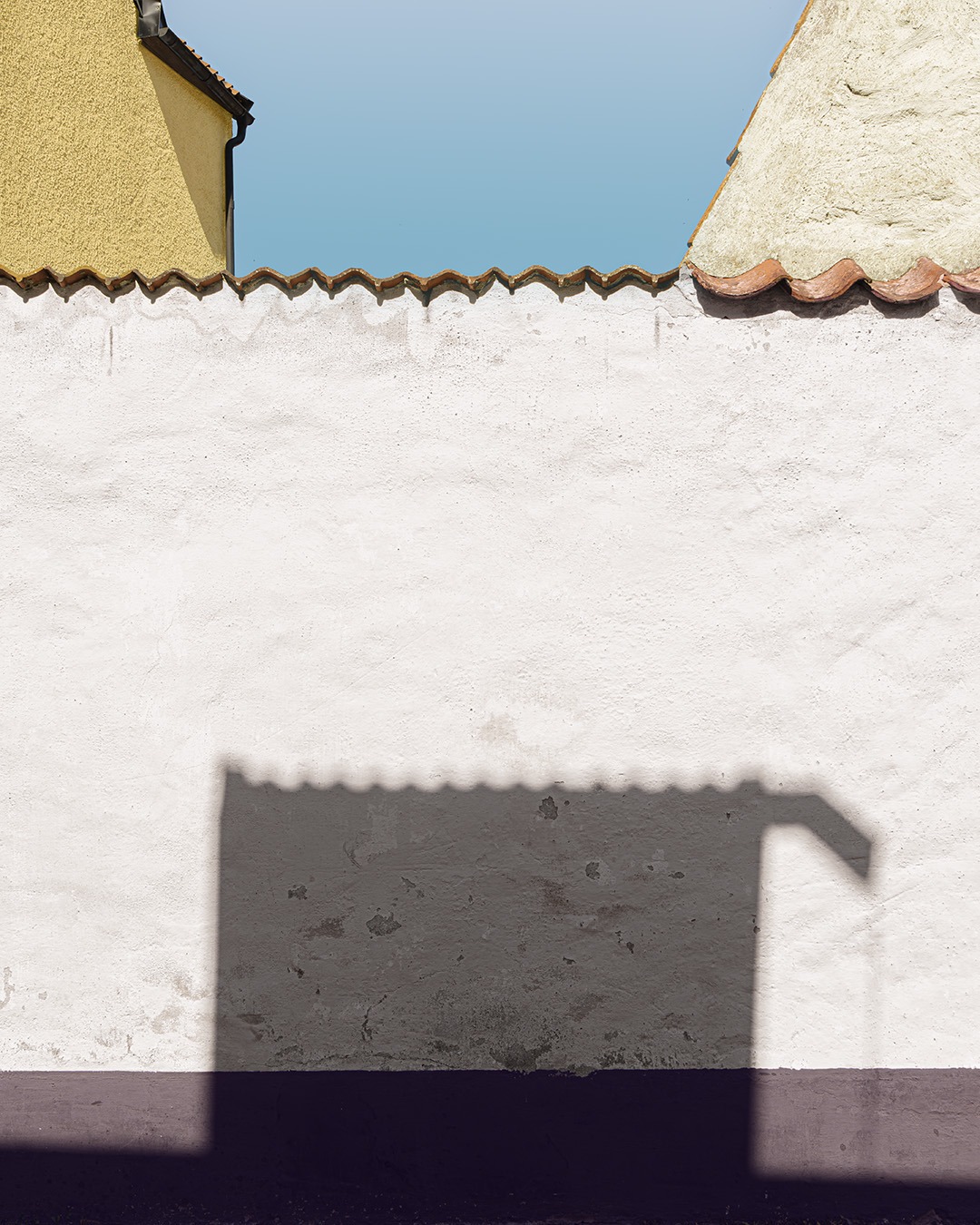 Colour-shifted fa&ccedil;ade study in Visby, Gotland: the shadow of a pantiled roof lying flat across a white rendered wall between an ochre and a cream gable, a dark violet plinth below.