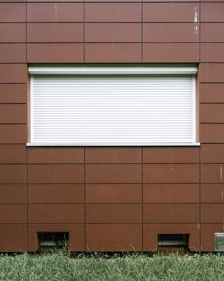 Red panelled facade with visible joints, a closed white roller shutter, two basement vents and a strip of grass below