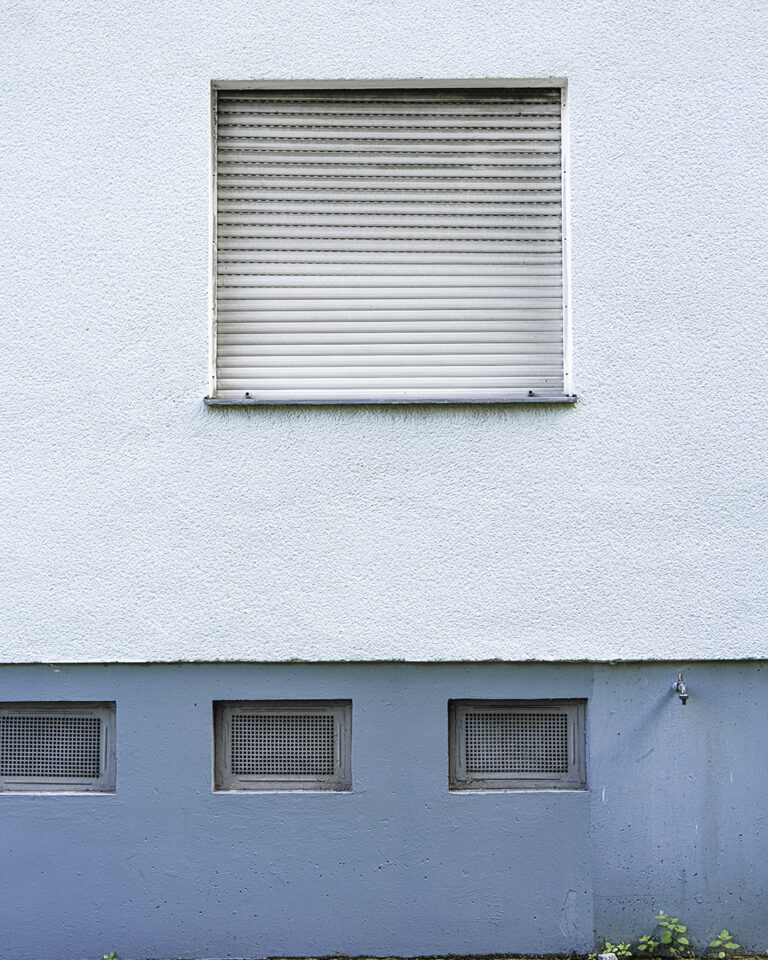Pale blue wall with a closed roller shutter above a stronger blue plinth carrying three barred basement windows
