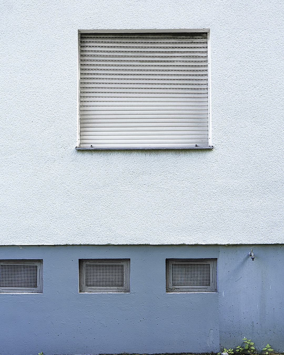Pale blue wall with a closed roller shutter above a stronger blue plinth carrying three barred basement windows