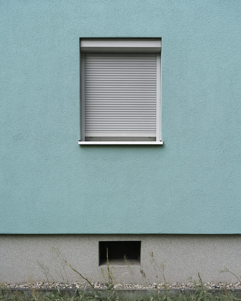 Mint green rendered wall with a single window and the white roller shutter fully down, a grey plinth with one basement vent, a gravel strip and weeds below