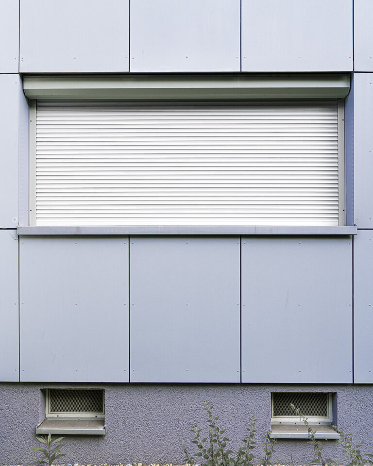 Grey-blue panelled facade with visible joints and a wide horizontal window, the white roller shutter fully down above a metal sill, a plinth with two basement vents and weeds below