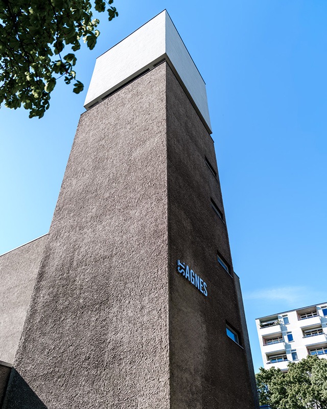The bell tower of St. Agnes from below against a deep blue sky, rough grey render lit from the side, a tree branch entering the frame at the top left