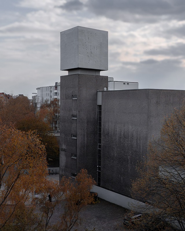 St. Agnes church in Berlin-Kreuzberg under an overcast sky: the grey rendered nave and the freestanding tower with its white top, seen across autumn trees